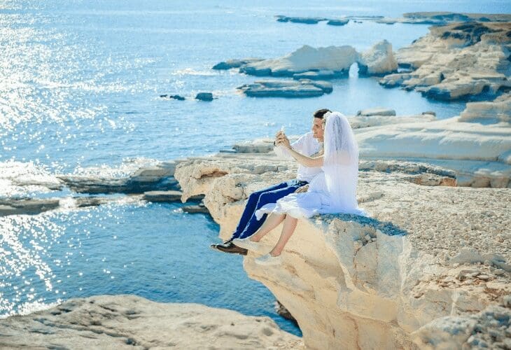 Brudepar sitter på et berg og tar bilder mot blått hav på en solfylt dag