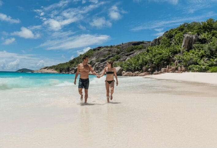 Et par går hånd i hånd på en strand i syden ikledd badetøy på en solfylt dag