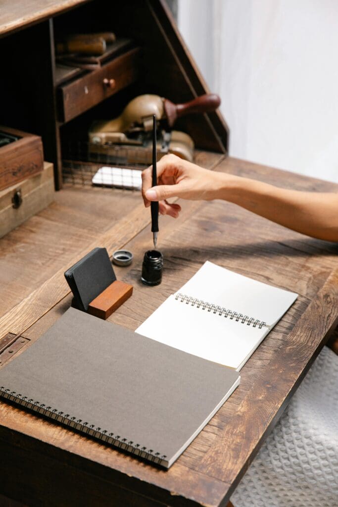 En hånd dypper en kalligrafipenn i blekk, klar til å skrive på et ark på en gammeldags kommode