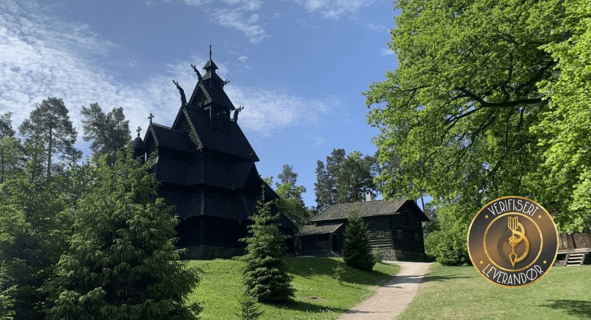 Sort stavkirke på Norsk folkemuseum mot grønn plen med grantrær