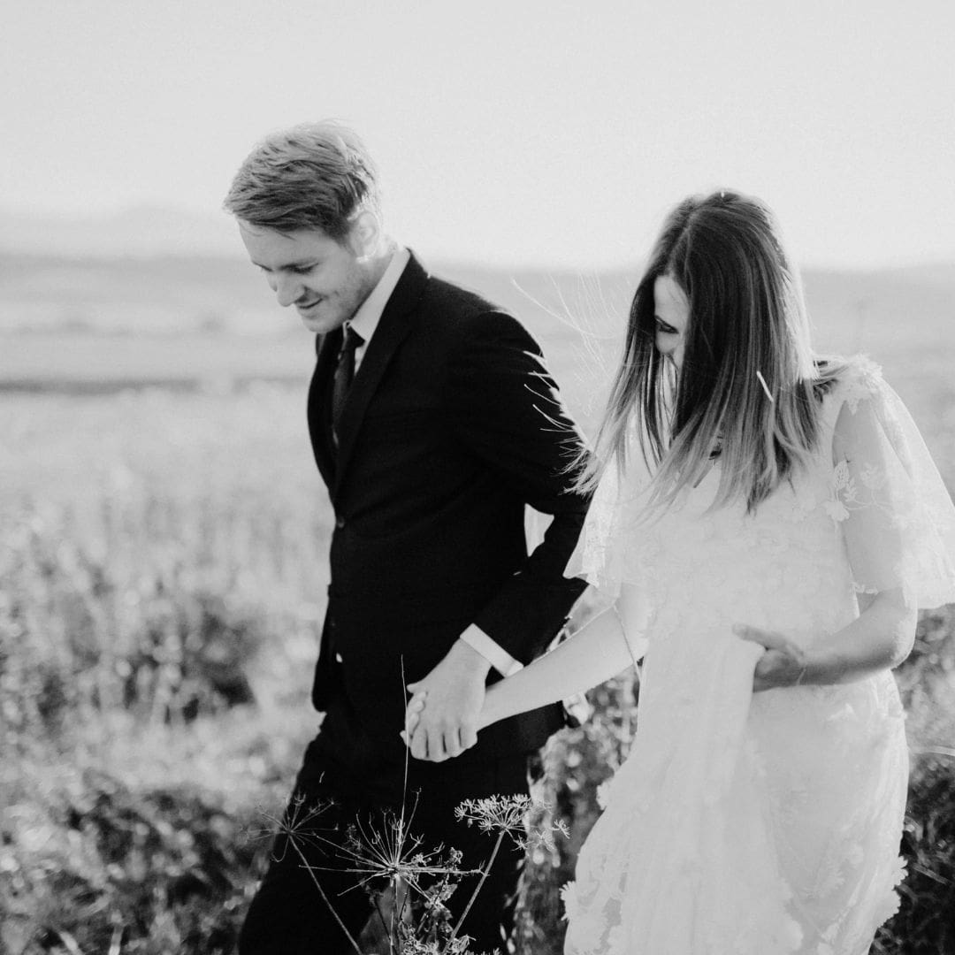 Et brudepar går hånd i hånd gjennom en åker med høyt gress
