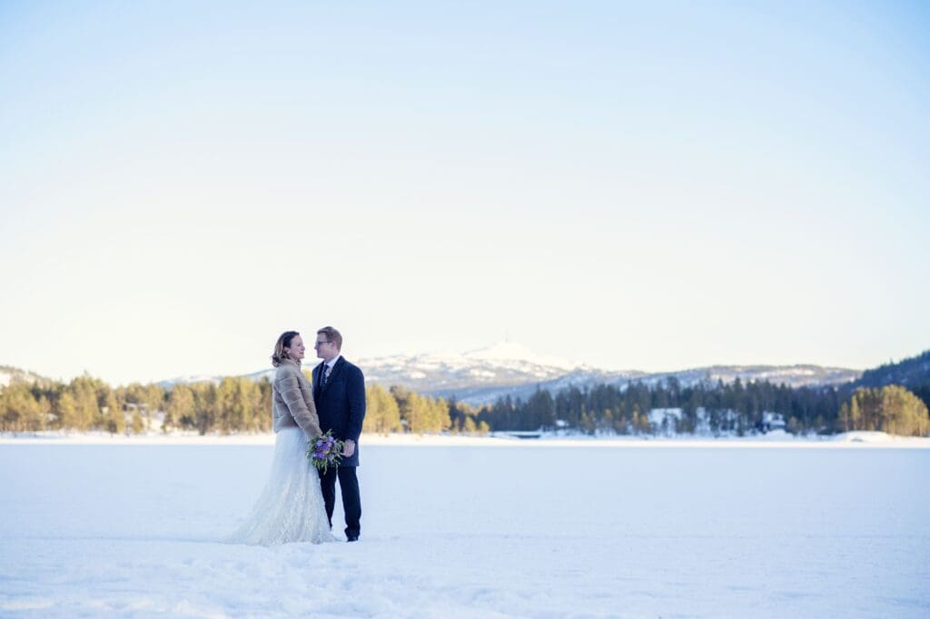 Brudepar holder hender under et vinterbryllup på en frossen innsjø dekket av snø mot lys blå himmel