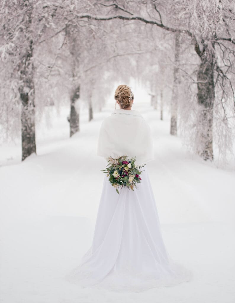Brud i vinterlandskap med kort hvit cape og brudebukett