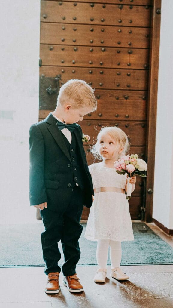 En gutt på ca 5 år, og en jente på ca 3 år står ved utgangen i en kirke, og representerer barn i bryllup. De er begge kledd i penklær. Barn under vielsen i et bryllup.