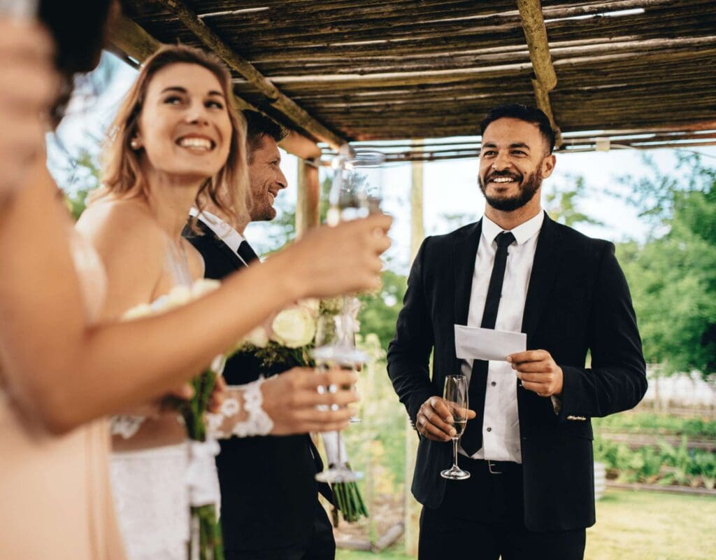 Mann i dress som holder et taleark og får gjestene til å le, med tre personer rundt ham som ser på. Toastmasteren som takler rollen.