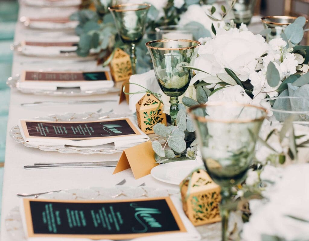 Elegant bordsetting til bryllup med menykort, sett fra siden, med flere tallerkner og bestikk synlige lenger bak i bildet.