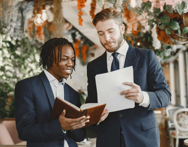 Toastmaster og taler i svart dress planlegger bryllupstaler med bryllupsblomster i bakgrunnen, den ene med ark og den andre med bok.