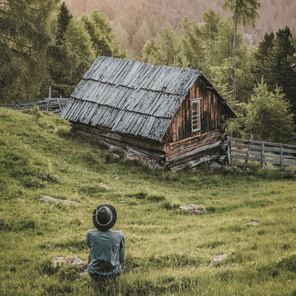 en person sitter med ryggen til i en dal, og ser på en trehytte. Personen har på seg hatt, og nyter stillheten. Representerer flukt fra den store oppgaven med bordplassering.