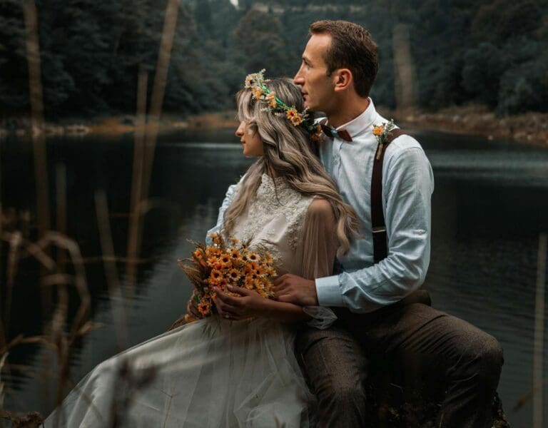 Et brudepar kledd i bryllupsklær sitter ved en innsjø, omringet av natur. Bruden har en blomsterkrans og de sitter med ryggen til kameraet, vendt mot sjøen.