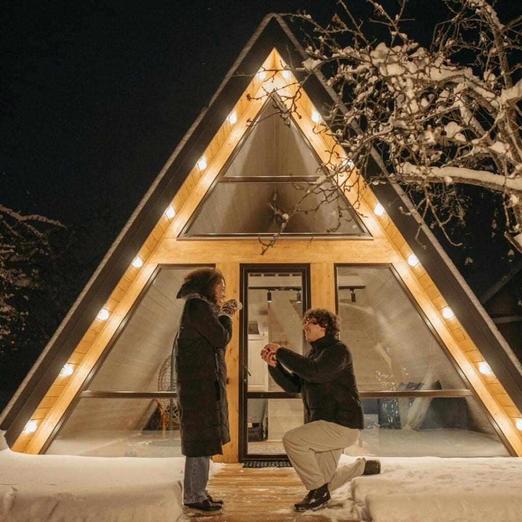 en mann frir til en kvinne utenfor en trekatet hytte, ute i snøfylt landskap