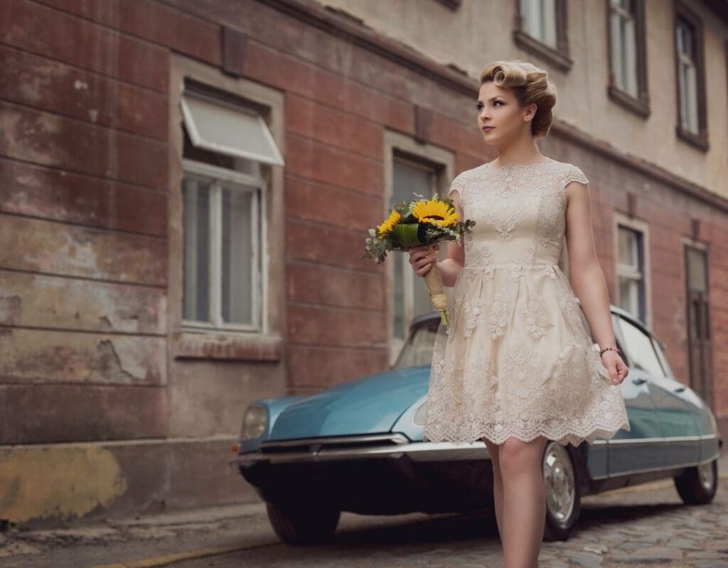 En retroinspirert brud i kort brudekjole med bukett står foran en vintage blå Porsche.